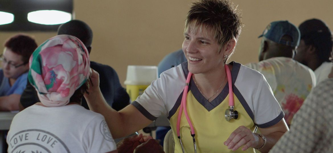 A smiling healthcare worker with a pink stethoscope gently interacts with a seated patient